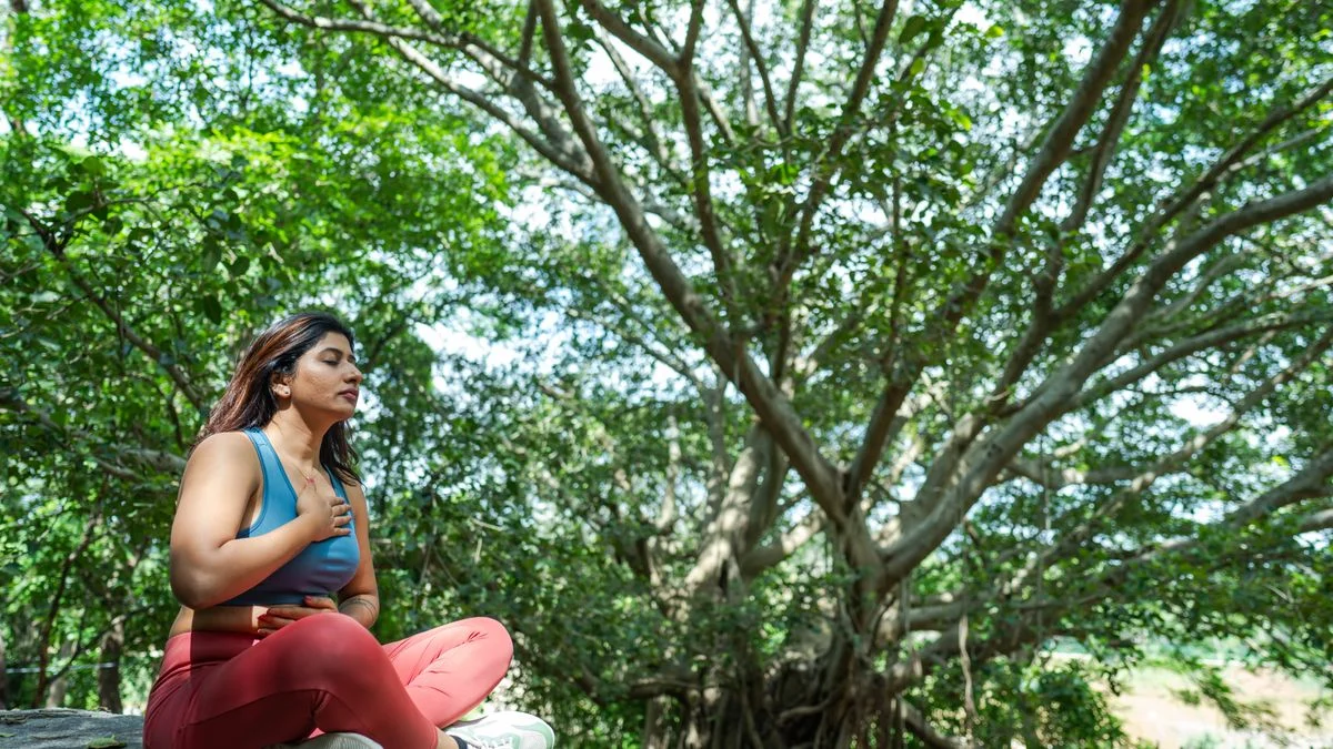 A woman sitting cross-legged outdoors under a large tree practising slow breathing
